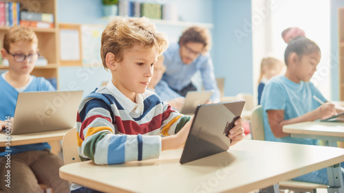 Elementary School Computer Science Class: Smart Boy Uses Digital Tablet Computer, His Classmates work with Laptops too. Children Getting Modern Education in STEM, Playing and Learning