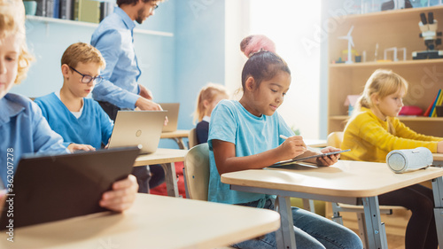 Elementary School Computer Science Class: Cute Girl Uses Digital Tablet Computer, Her Classmates work with Laptops too. Children Getting Modern Education in STEM, Playing and Learning