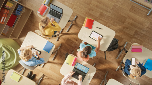 Top View Shot in Elementary School Computer Science Classroom: Children Sitting at their School Desk Using Personal Computers and Digital Tablets for Assignments. photo