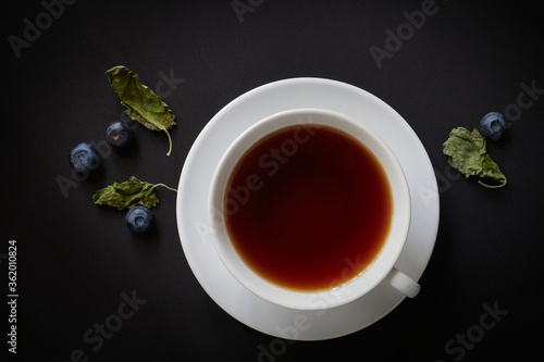 White cup with tea, blueberries and dried mint leaves on a dark background, view from above
