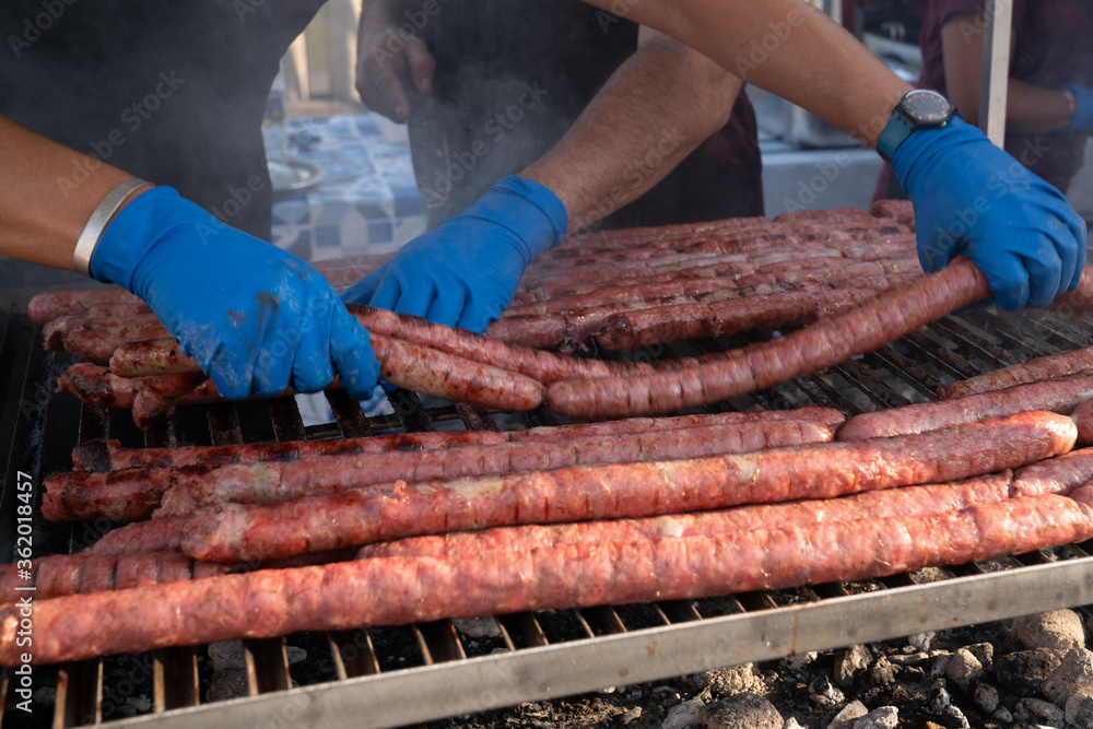 BBQ Sausage lined up on a large outdoor grill with smoke coming up from the fire below. person in black apron blue shirt gloved hand holding tongs turning meat