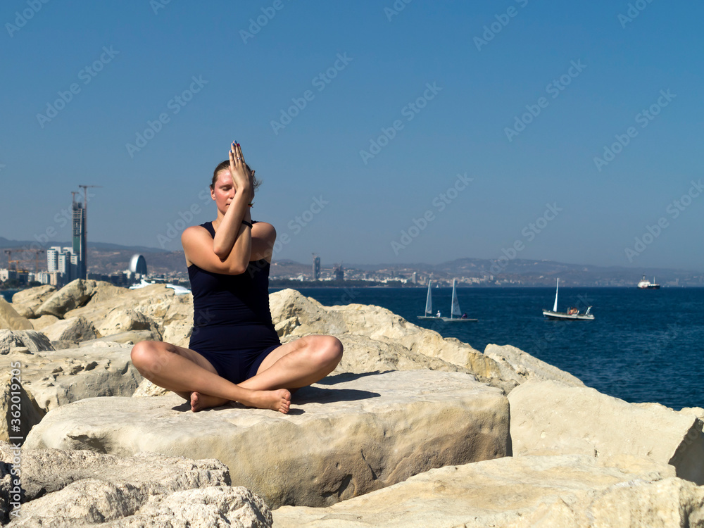young woman sitting cross legged on large rock by the seaside and practicing yoga 