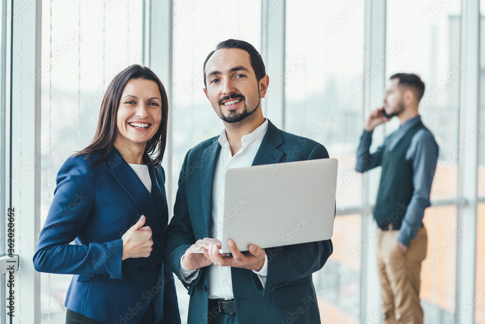 Successful man and woman making business plan, comparing annual data in laptop, while their coleague is on phone on blurred background. Successful man and woman making business plan, comparing annual data in laptop, while their coleague is on phone on blurred background.