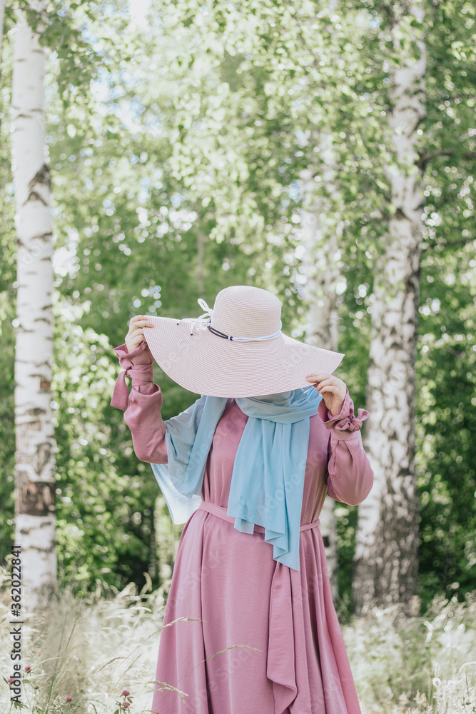 Muslim woman with a hat in a pink dress and a blue hijab with a picnic basket on a background of trees in summer. Modern muslim woman. Muslim woman in summer outfit. Ramadan. Beautiful muslim woman