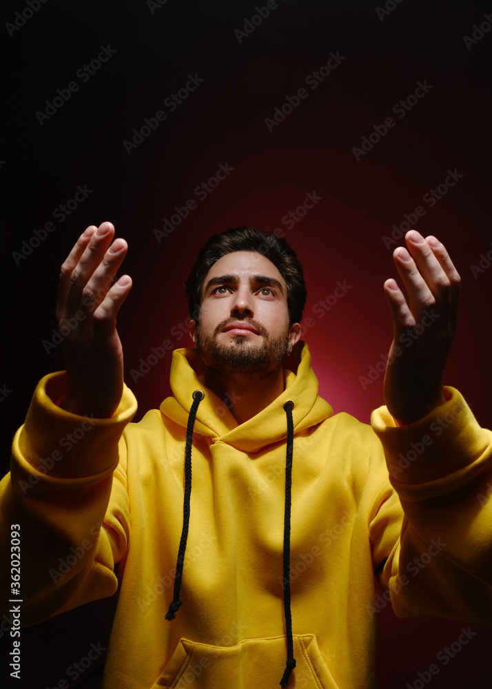 A young man of 25-30 years old in a yellow sweatshirt emotionally pulls his hands up on red wall background. 