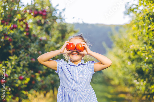 Cute smiling child in blue dress, holding apples in front of her eyes over blurred garden background.