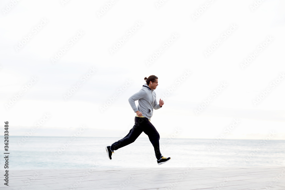 Sportive male runner working out outdoors jogging along seashore with beautiful sky horizon on background, jogger listening to music on smart phone while training outdoors