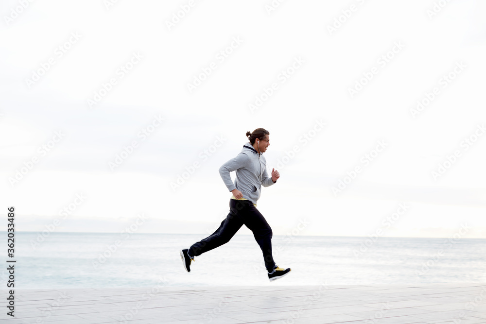 Sportive male runner working out outdoors jogging along seashore with beautiful sky horizon on background, jogger listening to music on smart phone while training outdoors Sportive male runner working out outdoors jogging along seashore with beautiful sky horizon on background, jogger listening to music on smart phone while training outdoors