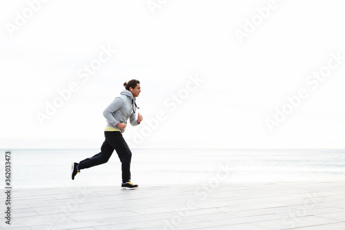 Young sportive man running along the beach listening to music on smart phone, athletic runner training on morning jog outdoors