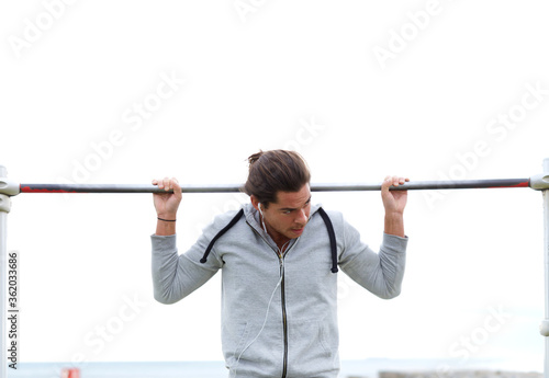 Half length portrait of young handsome man performs sport exercises to strengthen biceps muscles, male athlete doing pull ups on horizontal bar in a cloudy day while listening to music on headphones