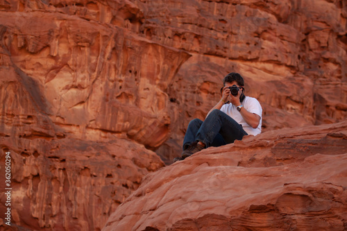 A European tourist visiting wadi rum is taking a photo of the marvelous desert landscape as he sits on top of a red sandstone rock. He wears jeans, white t-shirt and hiking shoes and holds a dslr.