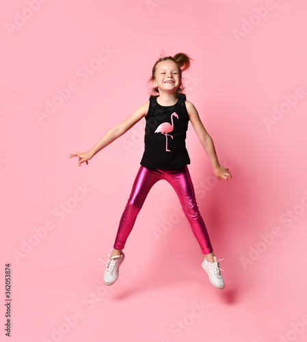Adorable little girl in sleeveless t-shirt with flamingo and bright rose leggings is merrily jumping on place. Full length shot isolated on pink