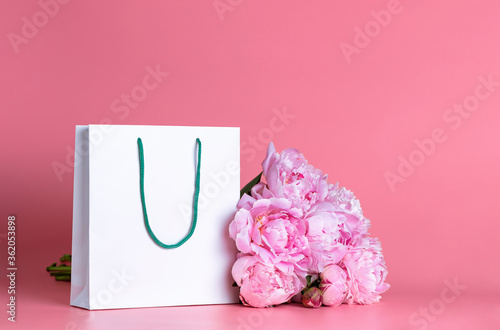 White gift bag with a bouquet of peonies on a pink background