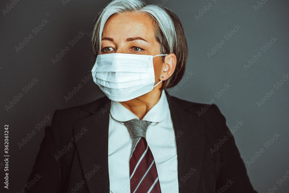 Gray-haired business woman wearing protective mask and office formalwear looking at side. Close up portrait on gray back. Tinted image. Gray-haired business woman wearing protective mask and office formalwear looking at side. Close up portrait on gray back. Tinted image.