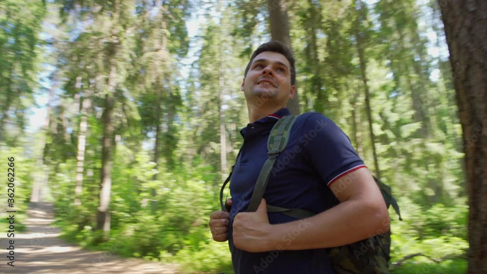 Tilt down panning shot of smiling middle aged adventure seeker with backpack standing among trees in forest and looking around admiring nature Tilt down panning shot of smiling middle aged adventure seeker with backpack standing among trees in forest and looking around admiring nature