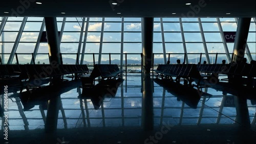 Time-lapse silhouette of unrecognizable people walking sitting in large room in airport where wait before going into a plane, blue cloudy sky view through the glass panoramic windows. Majorca, Spain