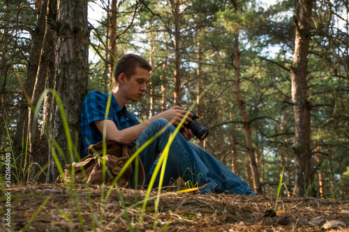 Young man in a blue plaid shirt with a camera in his hands in the coniferous forest