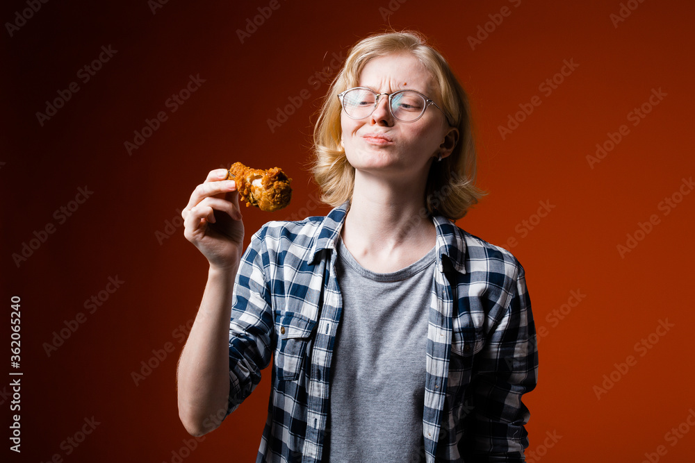 The blonde girl grimaces with displeasure and looks suspiciously at a chicken leg from fast food. The concept of rejecting junk food in favor of a healthy diet The blonde girl grimaces with displeasure and looks suspiciously at a chicken leg from fast food. The concept of rejecting junk food in favor of a healthy diet