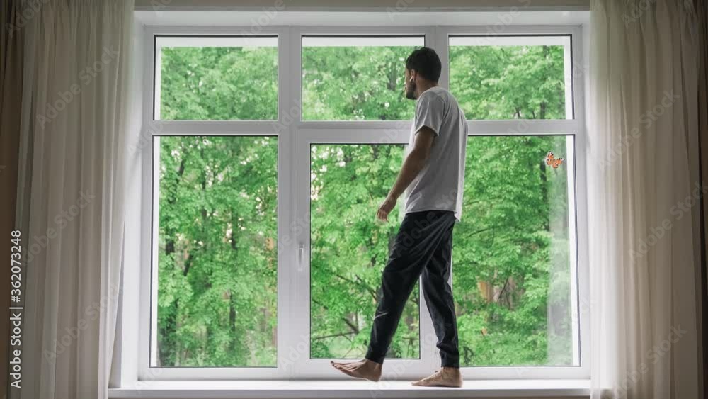 Corona virus. A self-isolated young sad man walks and stands, on the sill of a large window, looks far away and misses freedom. A person is at home alone during the quarantine, 4k Corona virus. A self-isolated young sad man walks and stands, on the sill of a large window, looks far away and misses freedom. A person is at home alone during the quarantine, 4k