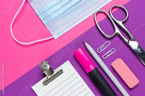 An empty checklist lies next to a medical mask, scissors, notebook, pen, eraser, paper clip and marker on a pink and purple background. Office work during the epidemic