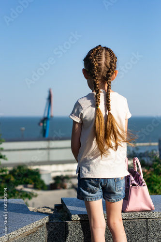 Little cute girl standing back near blue sea. Little girl from behind dressed in dress with white hat looking in to the sea. Baby standing on the sand and looking back near sea. Summer concept