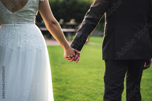 Rear view of married couple standing on lawn in back-light. Closeup of holding hands. Wedding day concept. © sebastiancaptures
