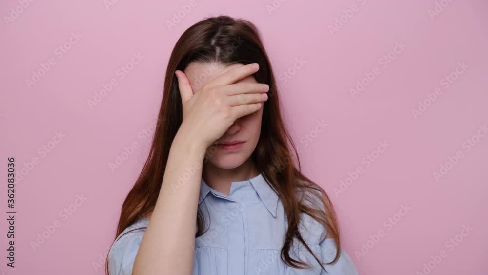 Portrait of disappointed young woman covers face with his hand, brunette girl feeling hopeless, suffering from stress and nervous tension, dressed in blue shirt, isolated on pink studio background Portrait of disappointed young woman covers face with his hand, brunette girl feeling hopeless, suffering from stress and nervous tension, dressed in blue shirt, isolated on pink studio background