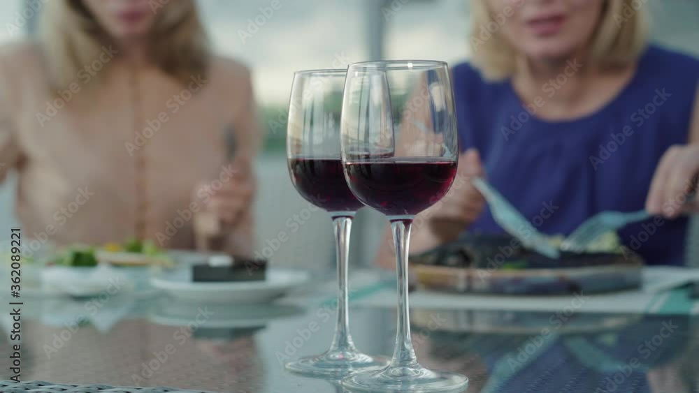 Two wineglasses with drink standing on the table as blurred women talking at the background. Expensive red wine in glasses ordered by wealthy female Caucasian clients in outdoor restaurant. Two wineglasses with drink standing on the table as blurred women talking at the background. Expensive red wine in glasses ordered by wealthy female Caucasian clients in outdoor restaurant.