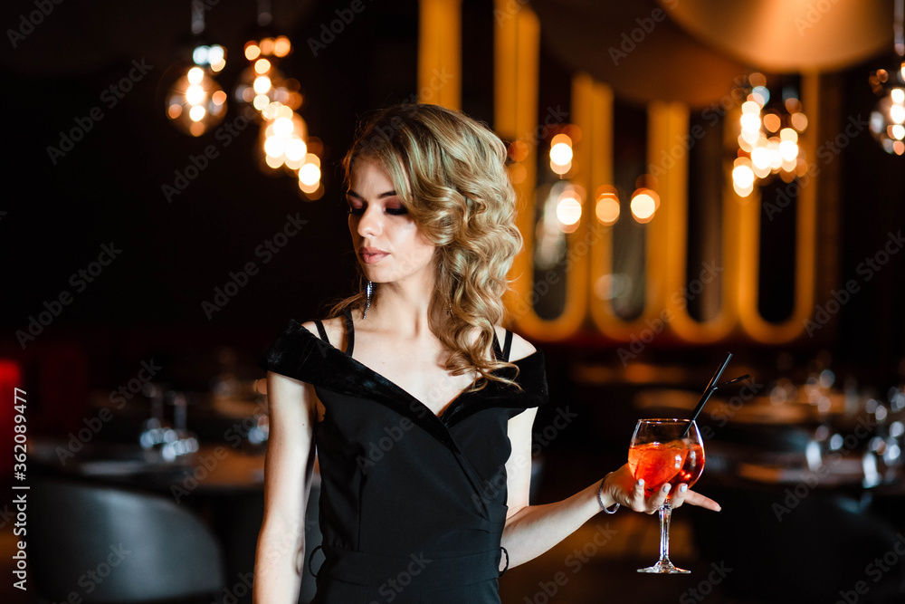 Beautiful blonde, young girl holding an aperol spritz. Cocktail aperol spritz in a glass. Portrait of a young girl in a restaurant. Portrait of a girl. Young girl in a restaurant with a glass. Beautiful blonde, young girl holding an aperol spritz. Cocktail aperol spritz in a glass. Portrait of a young girl in a restaurant. Portrait of a girl. Young girl in a restaurant with a glass.