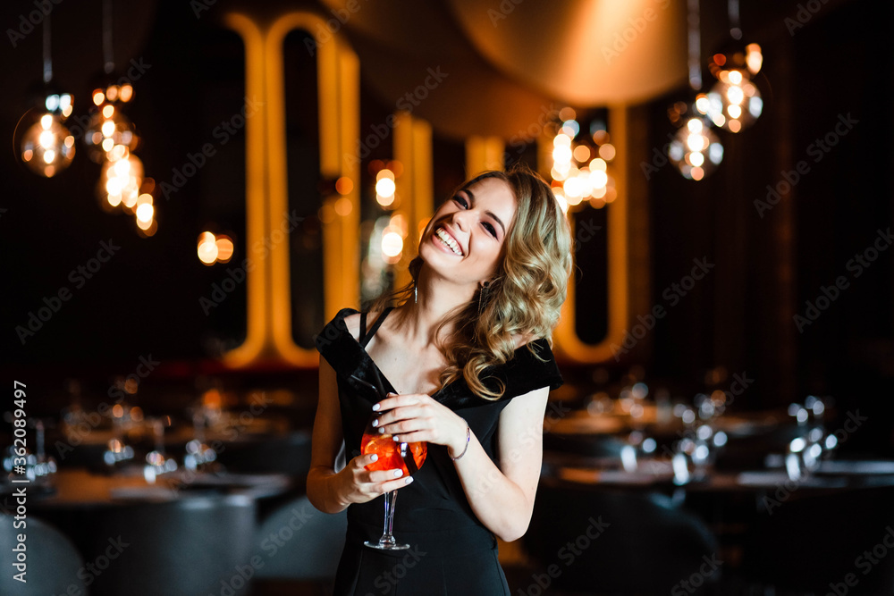 Beautiful blonde, young girl holding an aperol spritz. Cocktail aperol spritz in a glass. Portrait of a young girl in a restaurant. Portrait of a girl. Young girl in a restaurant with a glass. Beautiful blonde, young girl holding an aperol spritz. Cocktail aperol spritz in a glass. Portrait of a young girl in a restaurant. Portrait of a girl. Young girl in a restaurant with a glass.