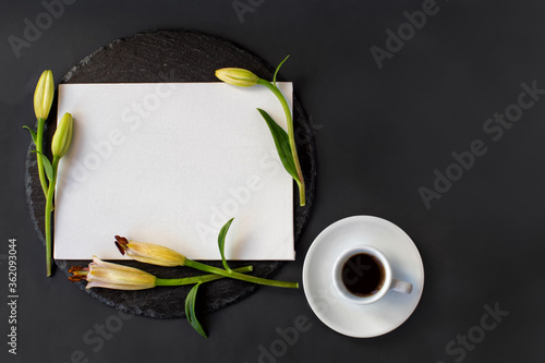 Cup of black coffee, white clean white textured banner paper card without massage with fresh spring buds of lily flowers on a round slate on a black background.  Top view. Space for text.