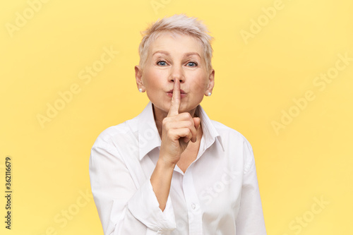Portrait of stylish attractive middle aged businesswoman in white shirt holding fore finger on her lips, asking you to keep silent about commercial secret, making shh gesture. Mature female shushing photo