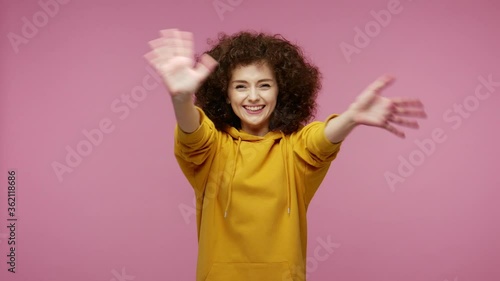 Sociable happy girl afro hairstyle in hoodie smiling friendly at camera and waving hands gesturing hello or goodbye, welcoming with hospitable expression. studio shot isolated on pink background photo