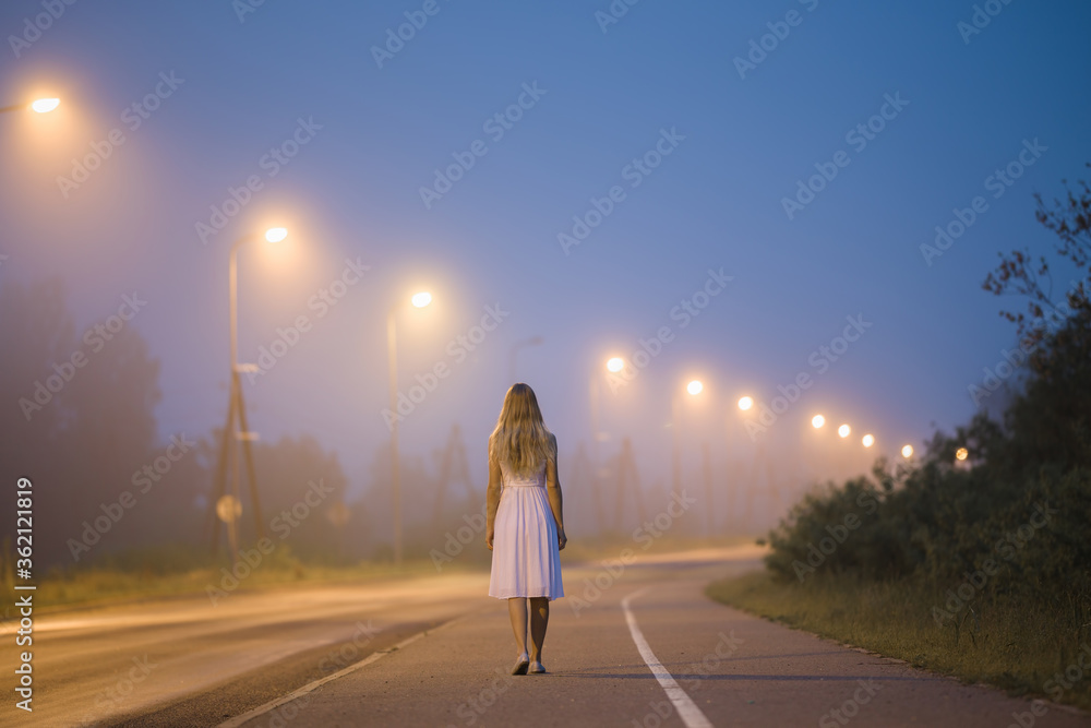 One young alone woman in white dress slowly walking on long sidewalk under street lights in dark summer night. Peaceful moment. Spending time alone. Back view.