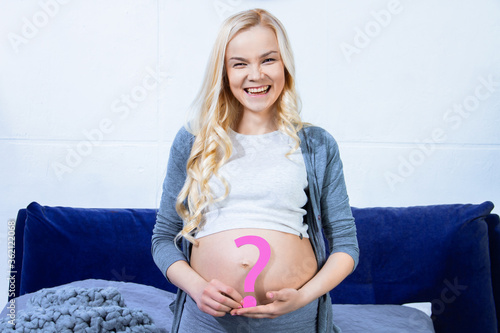 Young woman is holding pink paper question sign near abdomen, big pregnant female belly. Ultrasound diagnostics. Blonde parturient is expecting for little baby girl. Gender reveal party concept. photo