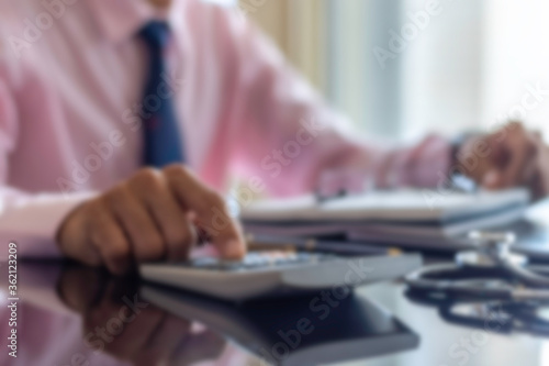 Blurred image of male doctor or physician calculating medicine costs with stethoscope and computer notebook on the office desk at clinic or hospital. Healthcare, medical costs and fees concept.