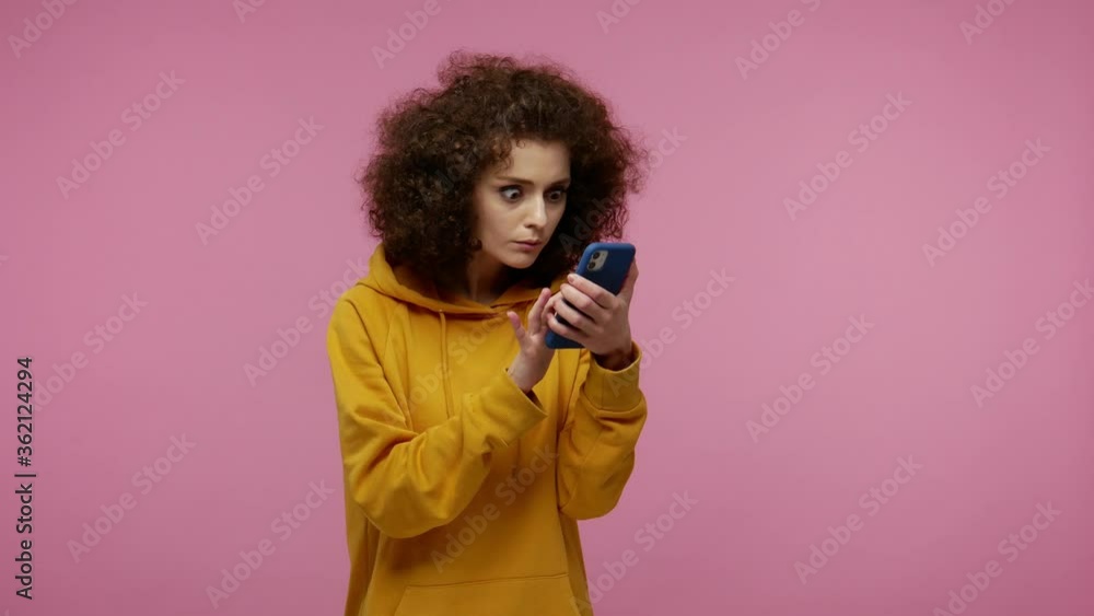 Girl afro hairstyle in hoodie scrolling social network using mobile phone and looking at camera with surprised expression, shocked by device or app. indoor studio shot isolated on pink background Girl afro hairstyle in hoodie scrolling social network using mobile phone and looking at camera with surprised expression, shocked by device or app. indoor studio shot isolated on pink background