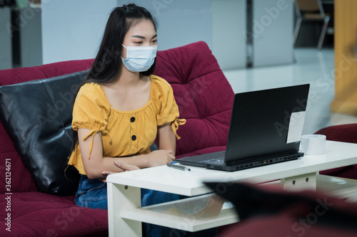 A teenager student wears surgical mask and studies online via laptop during COVID-19 pandemic. University student girl watches online classes and writing a syllabus in notebook. Stock photo.