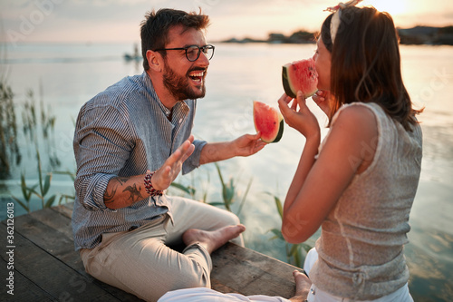 young adult couple sitting by the river at sunset, talking, laughing, eating watermelon