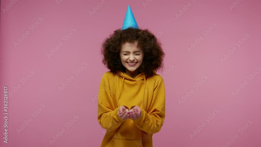 Optimistic girl afro hairstyle in hoodie with funny cone on her head laughing, moving in dance and blowing confetti glitters, celebrating birthday congratulating on anniversary. studio shot isolated Optimistic girl afro hairstyle in hoodie with funny cone on her head laughing, moving in dance and blowing confetti glitters, celebrating birthday congratulating on anniversary. studio shot isolated