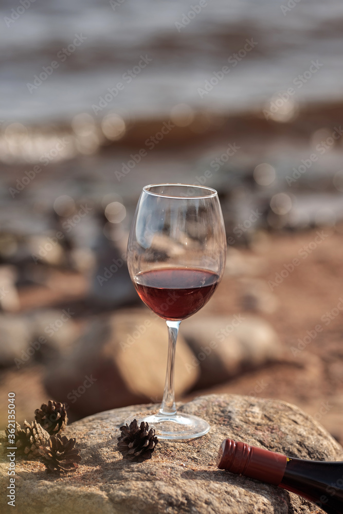 Red wine glass with bottle on the rocky sand. Summertime, vacation, weekend on Baltic sea, pine trees, northern nature