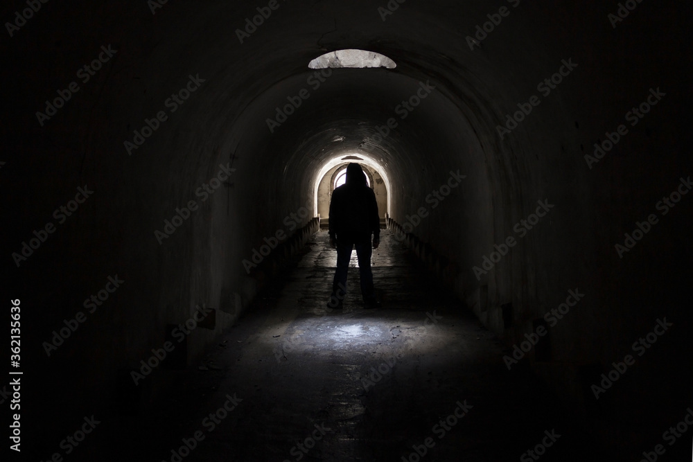 A silhouette of a man in dark clothes with a hood going to the exit to the lighted door from a dark terrible underground passage lit through a hatch in the ceiling.