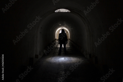 A silhouette of a man in dark clothes with a hood going to the exit to the lighted door from a dark terrible underground passage lit through a hatch in the ceiling. © Сергей Рамильцев