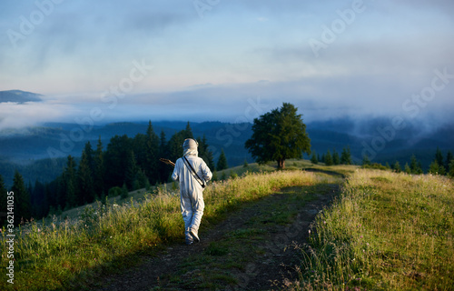 Back view of space traveler walking on mountain road with guitar. Cosmonaut guitarist in white space suit strolling down grassy trail through meadow with foggy hills and sky on background.