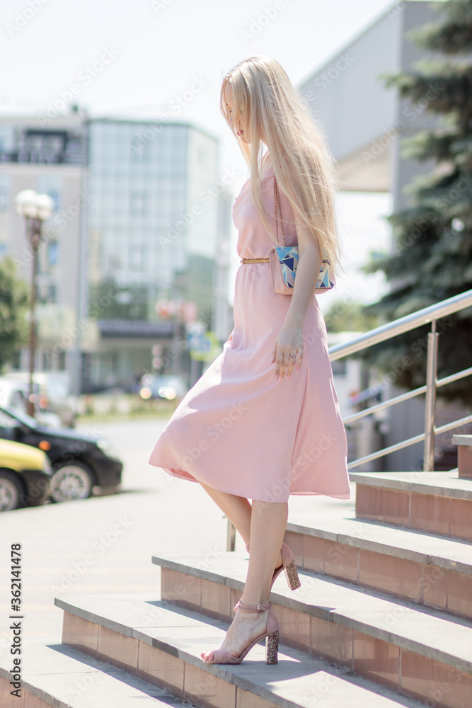 Fototapeta premium A young blonde girl, in a pink dress, descends the stairs of a shopping center on the street