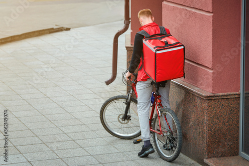 Courier on bicycle deliver food to costumer, wainting for online order and stands on bicycle on street. Man delivering food with red thermal backpack. Food delivery service photo