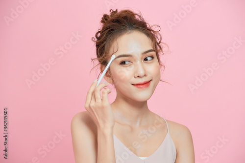 Portrait of excited young Asian woman holding brush and applying makeup over pink background.