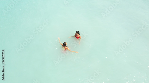 Aerial view of two young beautiful women in red bikini standing in turquoise azure blue water, swimming in sea ocean. Summer time holidays in tropics, vacation on Melasti beach Bali Indonesia Maldives