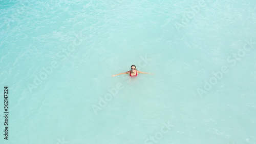 Aerial view of young beautiful teenager girl in red swimming suit sunglasses sit in light blue azure turquoise sea water, relaxing in ocean. Beach vacation. Malibu lifeguard. Maldives. Melasti beach.