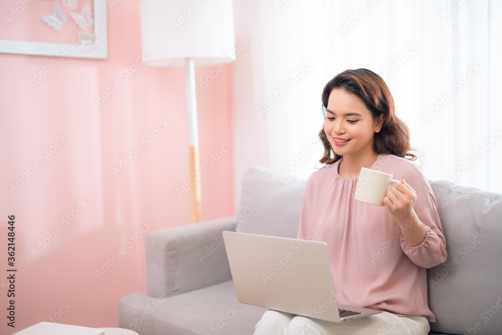 Attractive Asian female lifestyle using the laptop sitting on a couch and drinking coffee Attractive Asian female lifestyle using the laptop sitting on a couch and drinking coffee